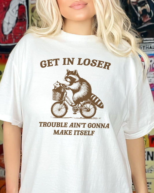 Woman wearing a white 'Get In Loser, Trouble Ain't Gonna Make Itself' t-shirt featuring a funny retro raccoon riding a bicycle graphic.
