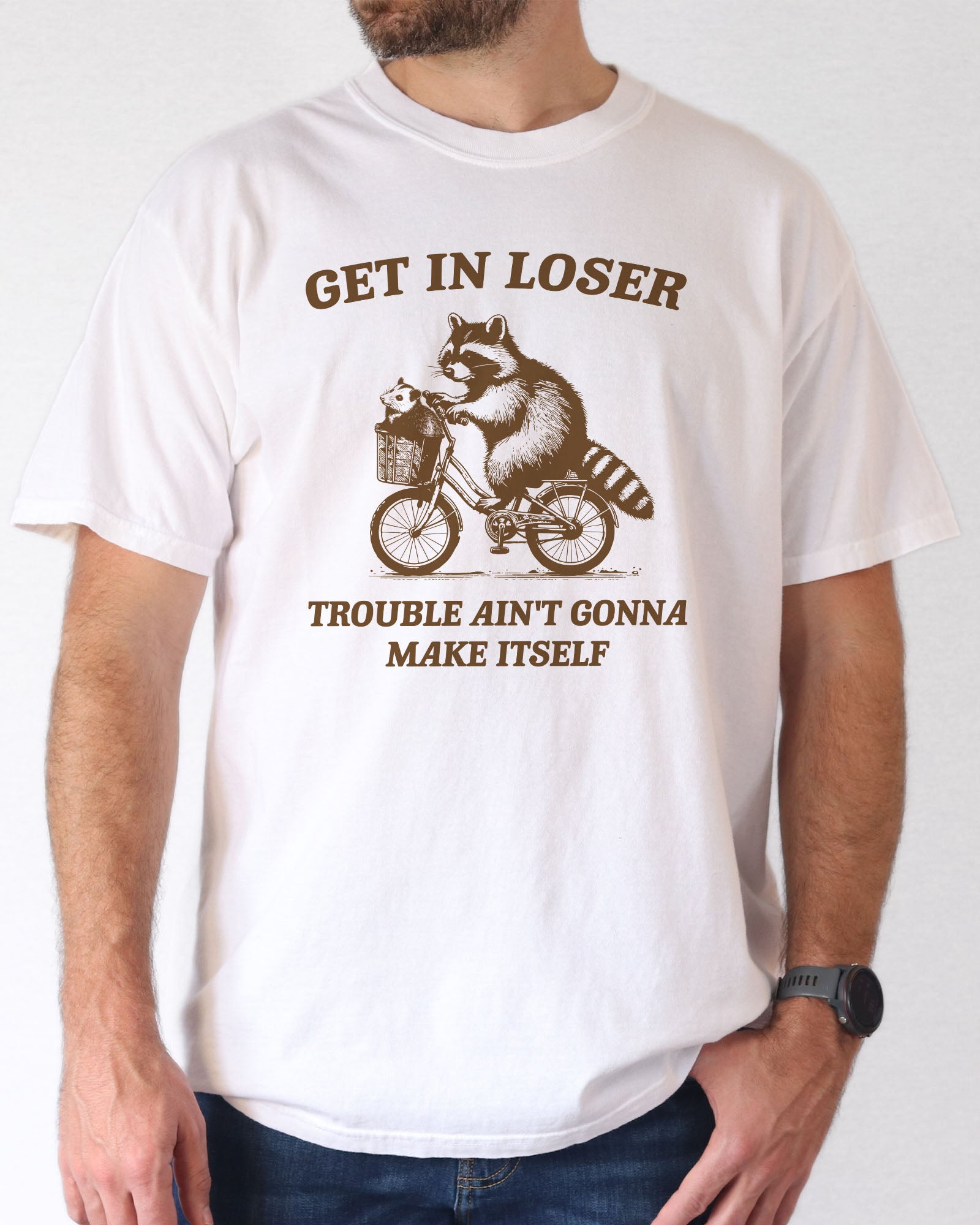 Man wearing a white color 'Get In Loser, Trouble Ain't Gonna Make Itself' graphic tee featuring a funny retro raccoon riding a bicycle graphic.