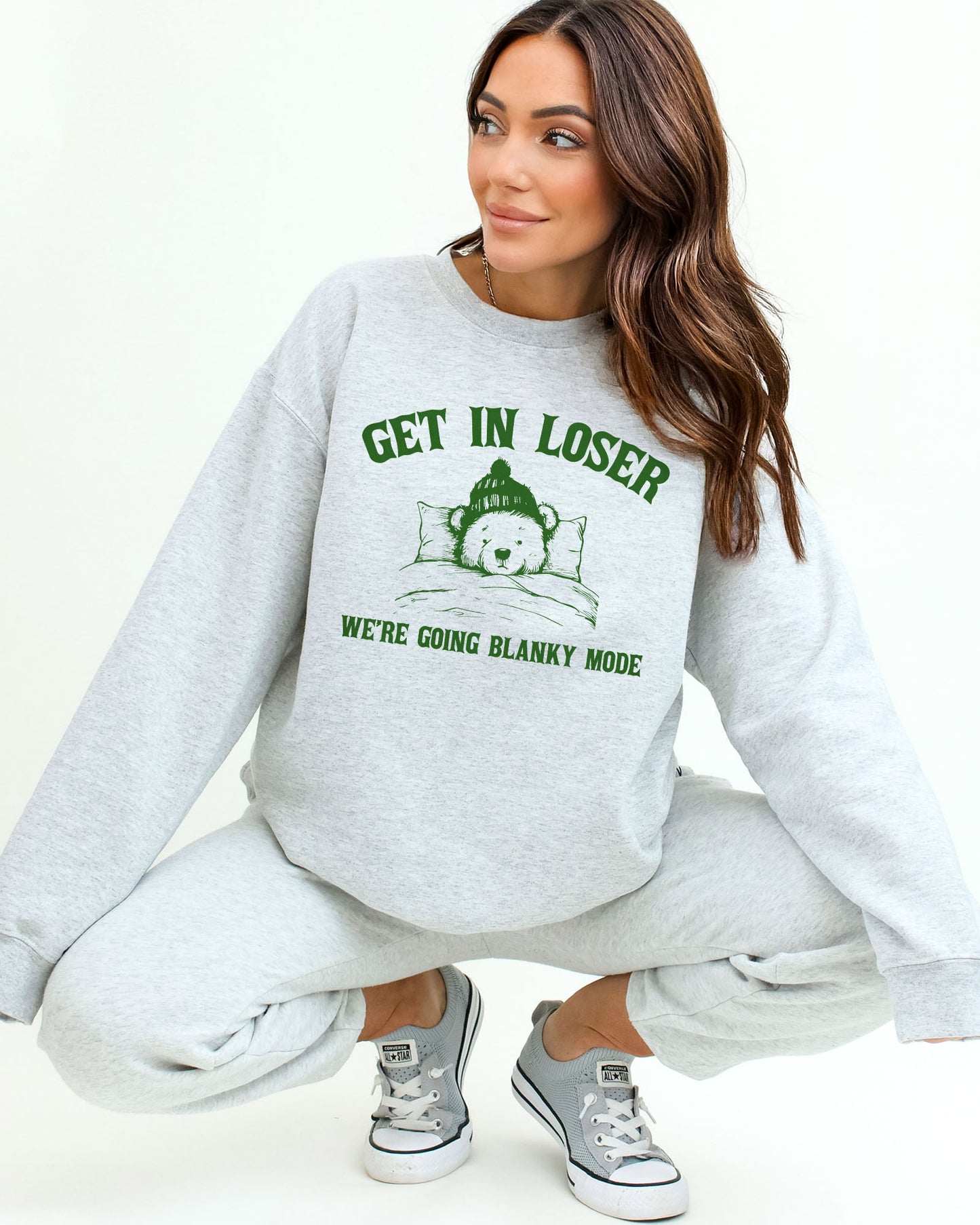 Woman wearing an ash gray sweatshirt with a funny retro graphic of a teddy bear in bed and the quote "Get In Loser We're Going Blanky Mode".