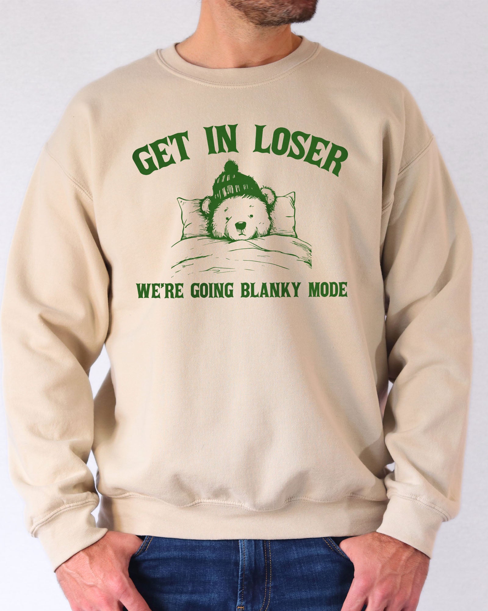 Man wearing a sand beige sweatshirt with a funny retro graphic of a teddy bear in bed and the quote "Get In Loser We're Going Blanky Mode".