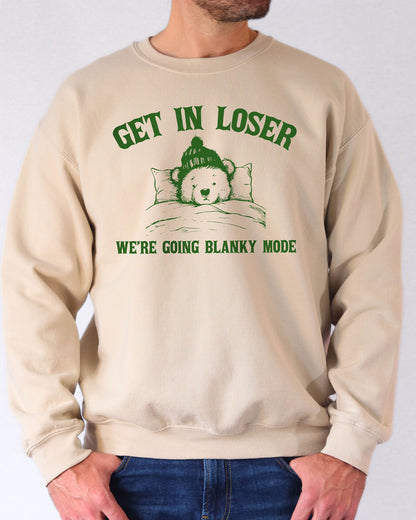 Man wearing a sand beige sweatshirt with a funny retro graphic of a teddy bear in bed and the quote "Get In Loser We're Going Blanky Mode".