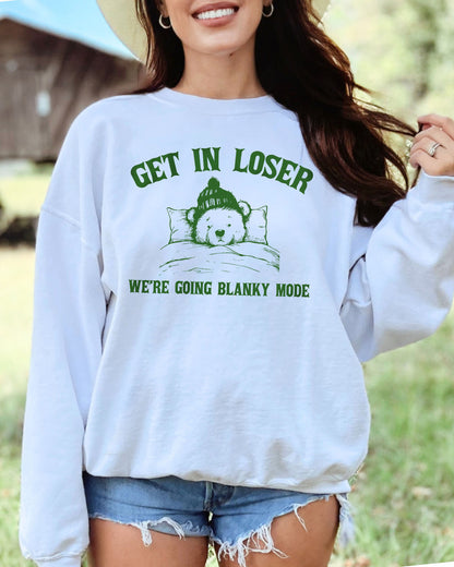 Woman wearing a white sweatshirt with a funny retro graphic of a teddy bear in bed and the quote "Get In Loser We're Going Blanky Mode".