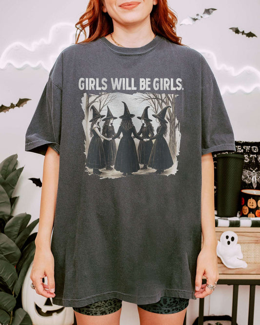 Woman wearing a pepper-colored “Girls Will Be Girls” shirt with a retro graphic of witches performing a witchy ritual.