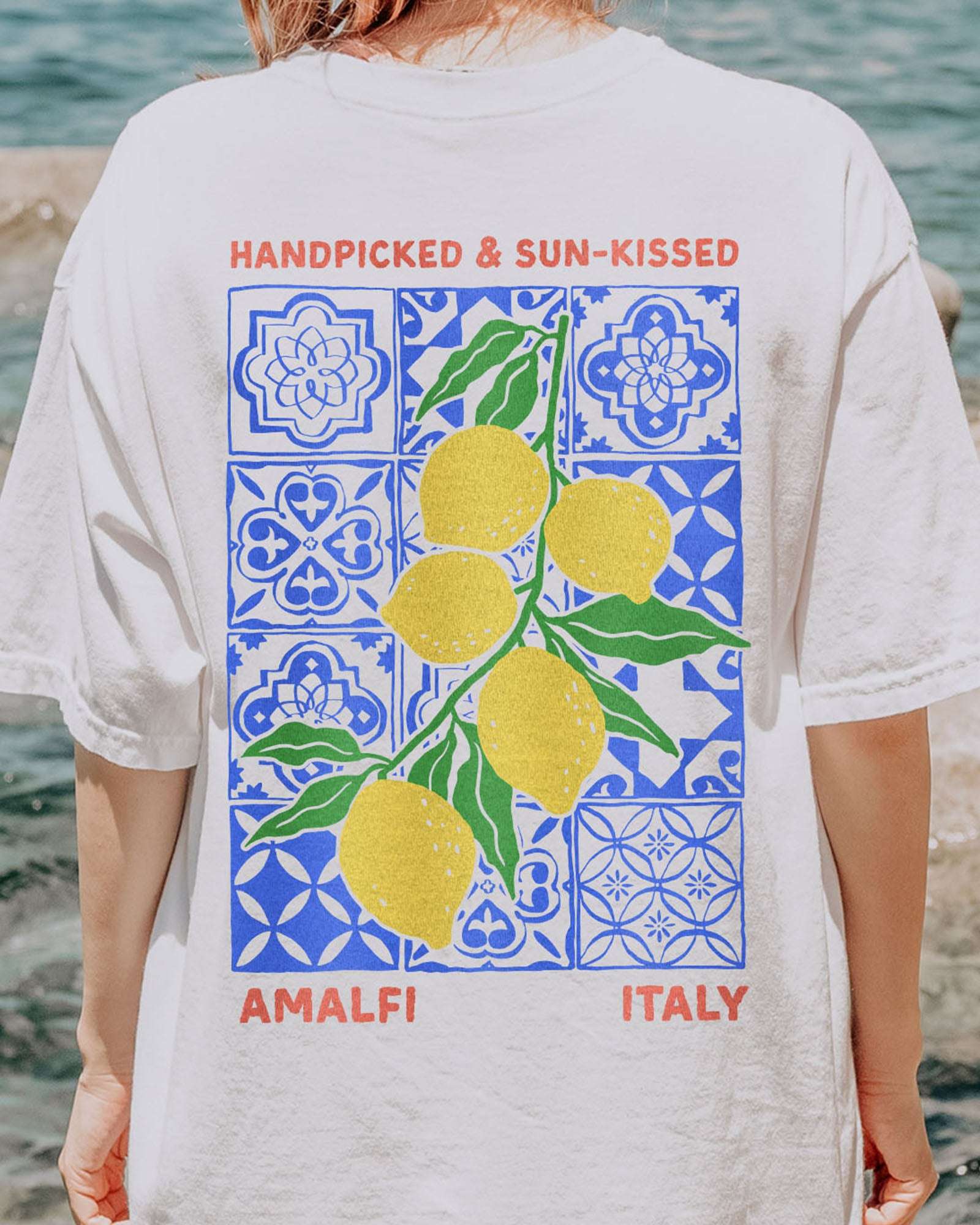 Close-up of woman wearing a white "Handpicked & Sun-Kissed Amalfi Italy" shirt with a retro graphic of lemons over a floral pattern background.