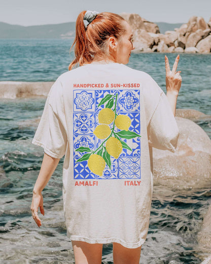 Woman wearing an ivory "Handpicked & Sun-Kissed Amalfi Italy" shirt with a retro graphic of lemons over a floral pattern background.