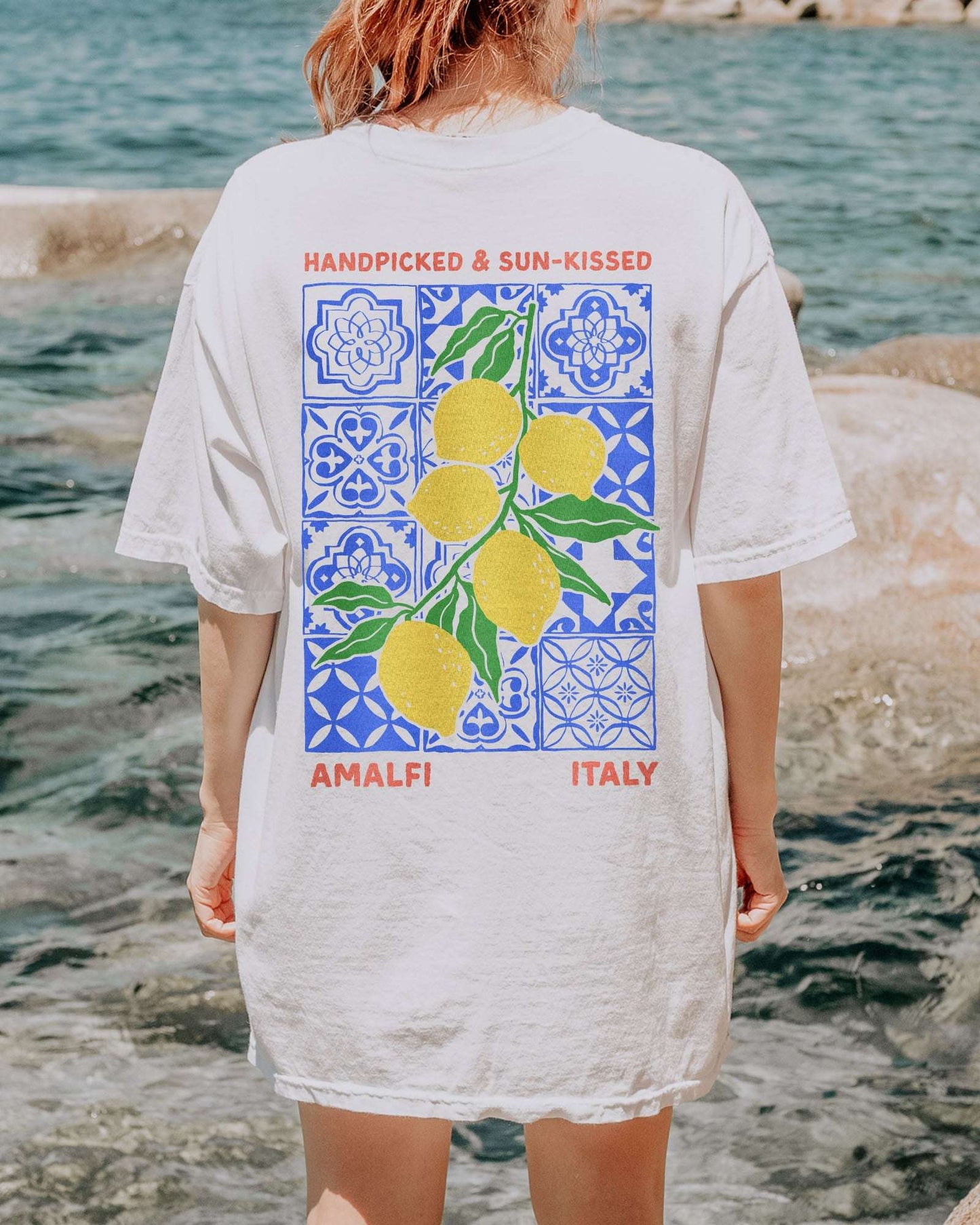 Woman wearing a white "Handpicked & Sun-Kissed Amalfi Italy" shirt with a retro graphic of lemons over a floral pattern background.