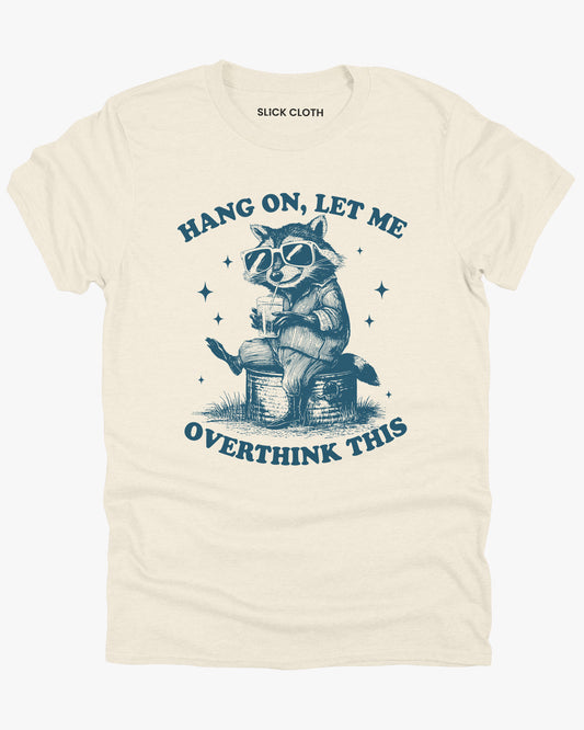 Flat lay natural beige 'Hang On Let Me Overthink This' shirt with funny retro raccoon wearing sunglasses graphic