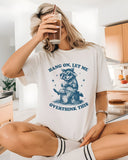 Woman sitting on kitchen counter and wearing white 'Hang On Let Me Overthink This' shirt with funny retro raccoon wearing sunglasses graphic.