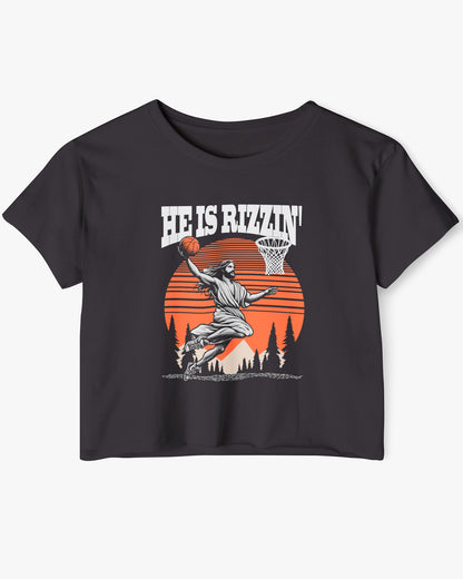 Flat lay of a black 'He Is Rizzin' crop top featuring a funny retro graphic of Jesus slam-dunking a basketball.