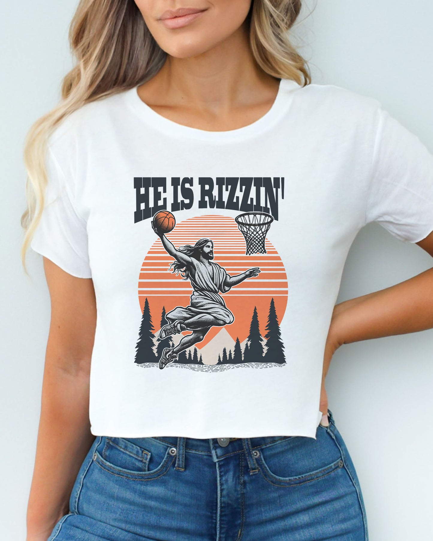 Woman wearing a white 'He Is Rizzin' crop top with a funny retro graphic of Jesus dunking a basketball.