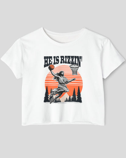 Flat lay of a white 'He Is Rizzin' crop top featuring a funny retro graphic of Jesus slam-dunking a basketball.