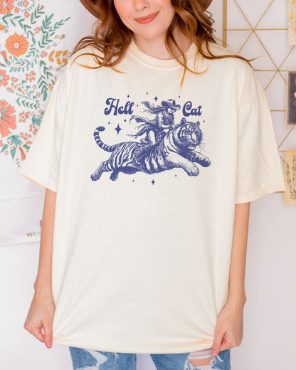 Woman wearing an ivory "Hell Cat" shirt with a retro graphic of a cowgirl riding a tiger.