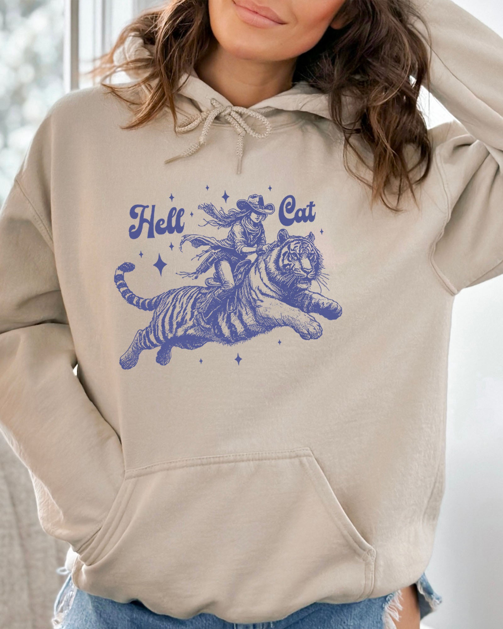 Woman wearing a sand-colored hoodie featuring a retro cowgirl riding a tiger graphic, titled 'Hell Cat'.