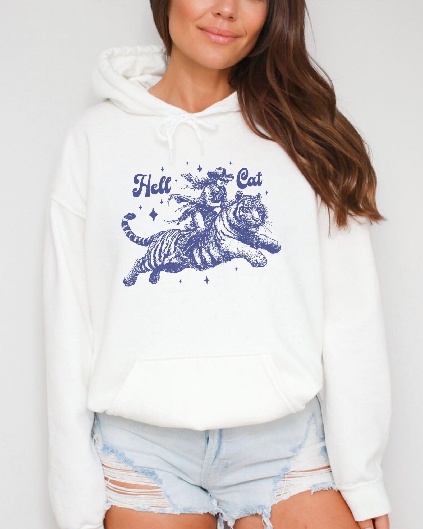 Woman wearing a white-colored hoodie featuring a retro cowgirl riding a tiger graphic, titled 'Hell Cat'.