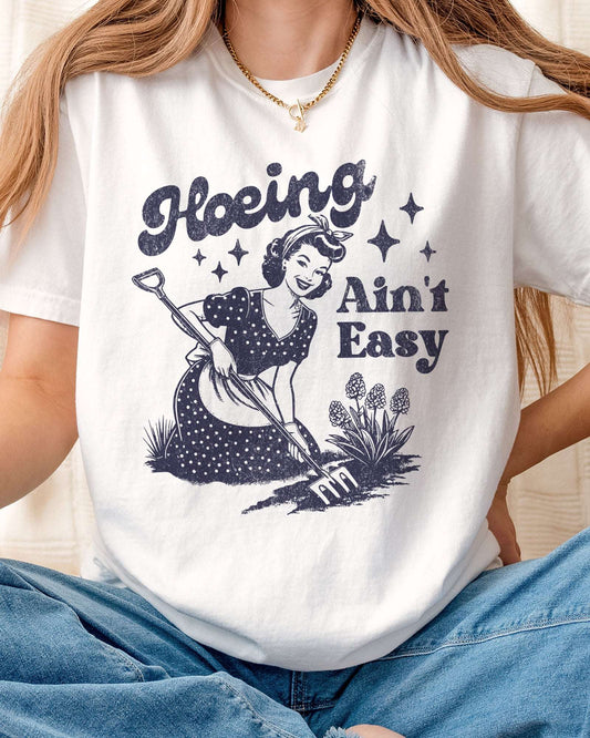 Close-up of "Hoeing Ain't Easy" shirt in white color with a funny retro graphic of a housewife in the garden.