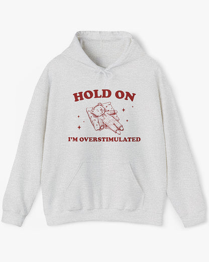 Flat lay ash gray 'Hold On I'm Overstimulated' hoodie with a retro and funny exhausted bear laying on pillow graphic