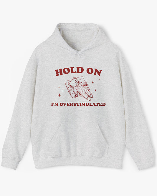 Flat lay ash gray 'Hold On I'm Overstimulated' hoodie with a retro and funny exhausted bear laying on pillow graphic