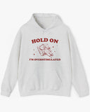 Flat lay ash gray 'Hold On I'm Overstimulated' hoodie with a retro and funny exhausted bear laying on pillow graphic