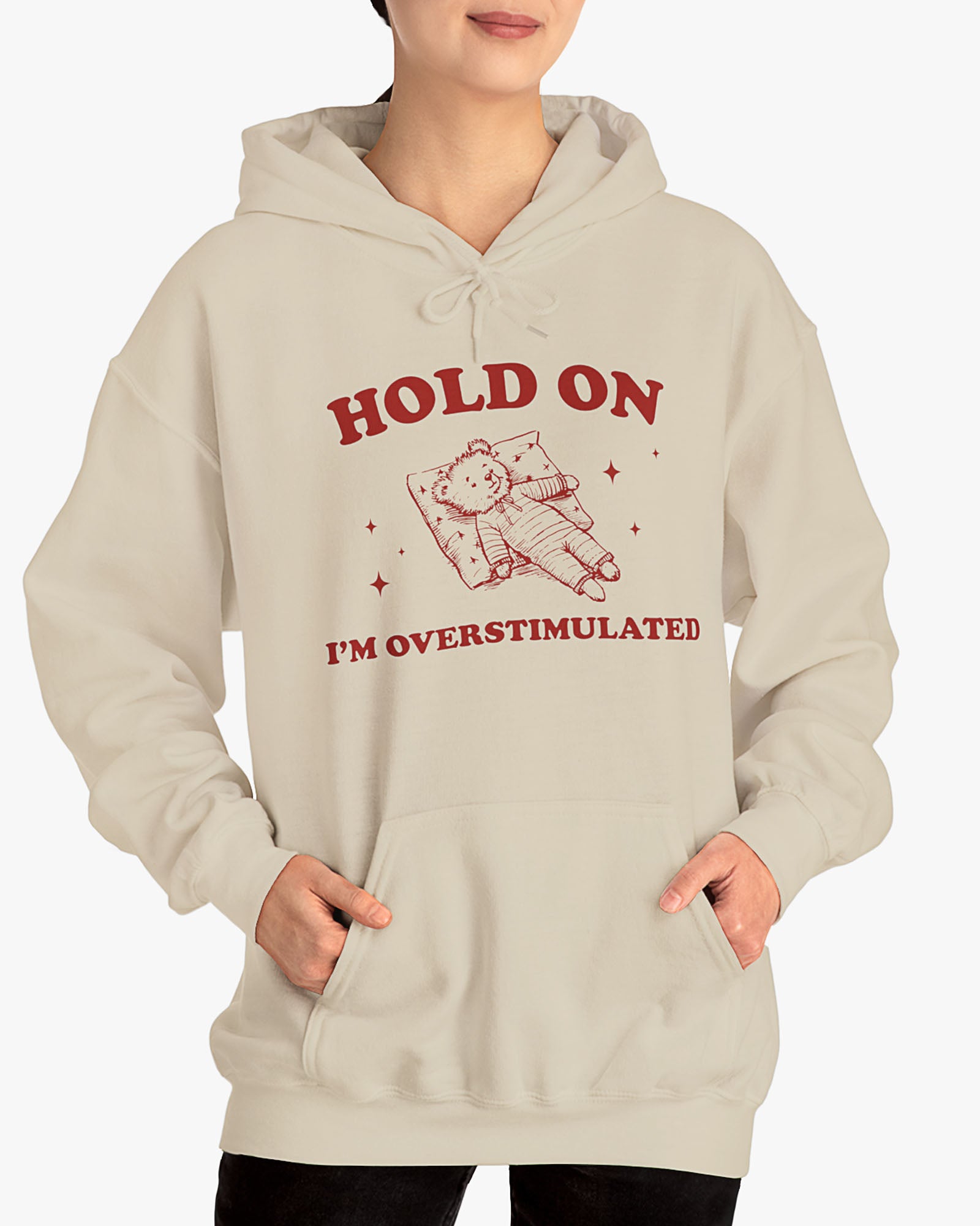 Woman wearing sand beige 'Hold On I'm Overstimulated' hoodie with a retro and funny exhausted bear laying on pillow graphic