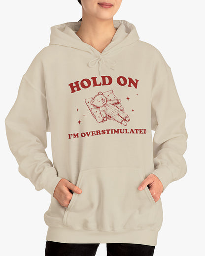 Woman wearing sand beige 'Hold On I'm Overstimulated' hoodie with a retro and funny exhausted bear laying on pillow graphic
