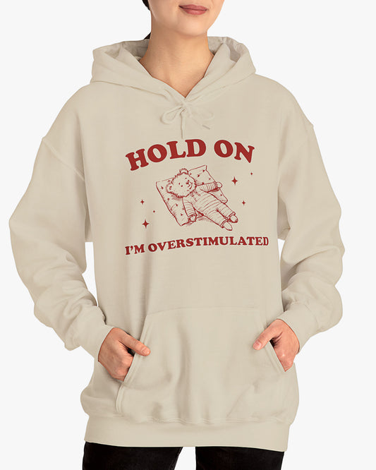 Woman wearing sand beige 'Hold On I'm Overstimulated' hoodie with a retro and funny exhausted bear laying on pillow graphic
