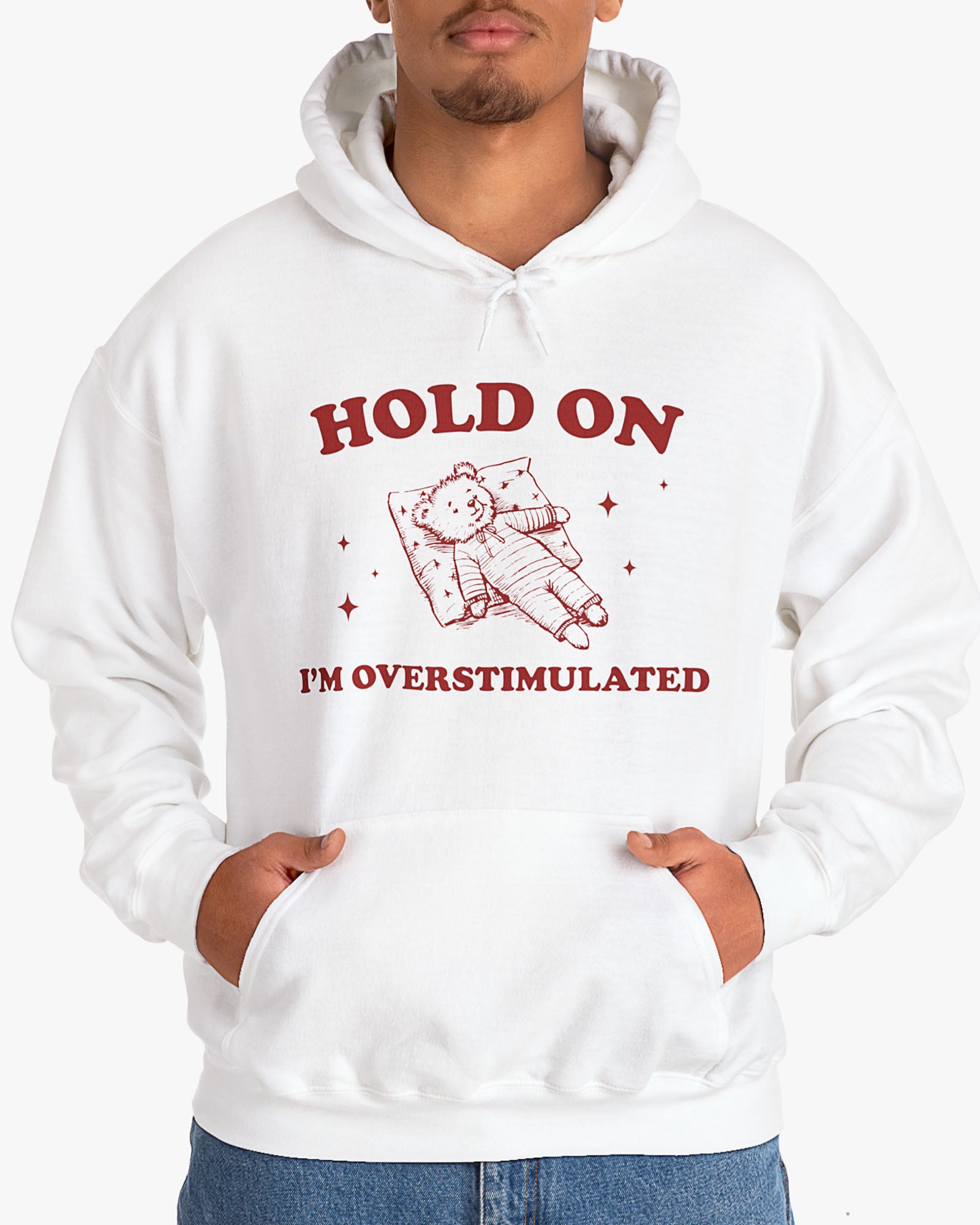 Man wearing white 'Hold On I'm Overstimulated' hoodie with a retro and funny exhausted bear laying on pillow graphic