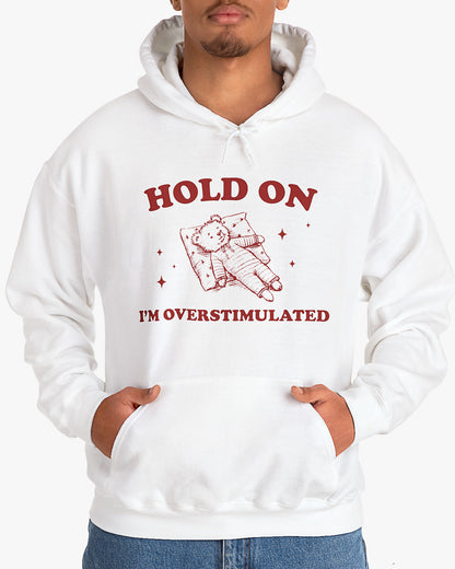 Man wearing white 'Hold On I'm Overstimulated' hoodie with a retro and funny exhausted bear laying on pillow graphic