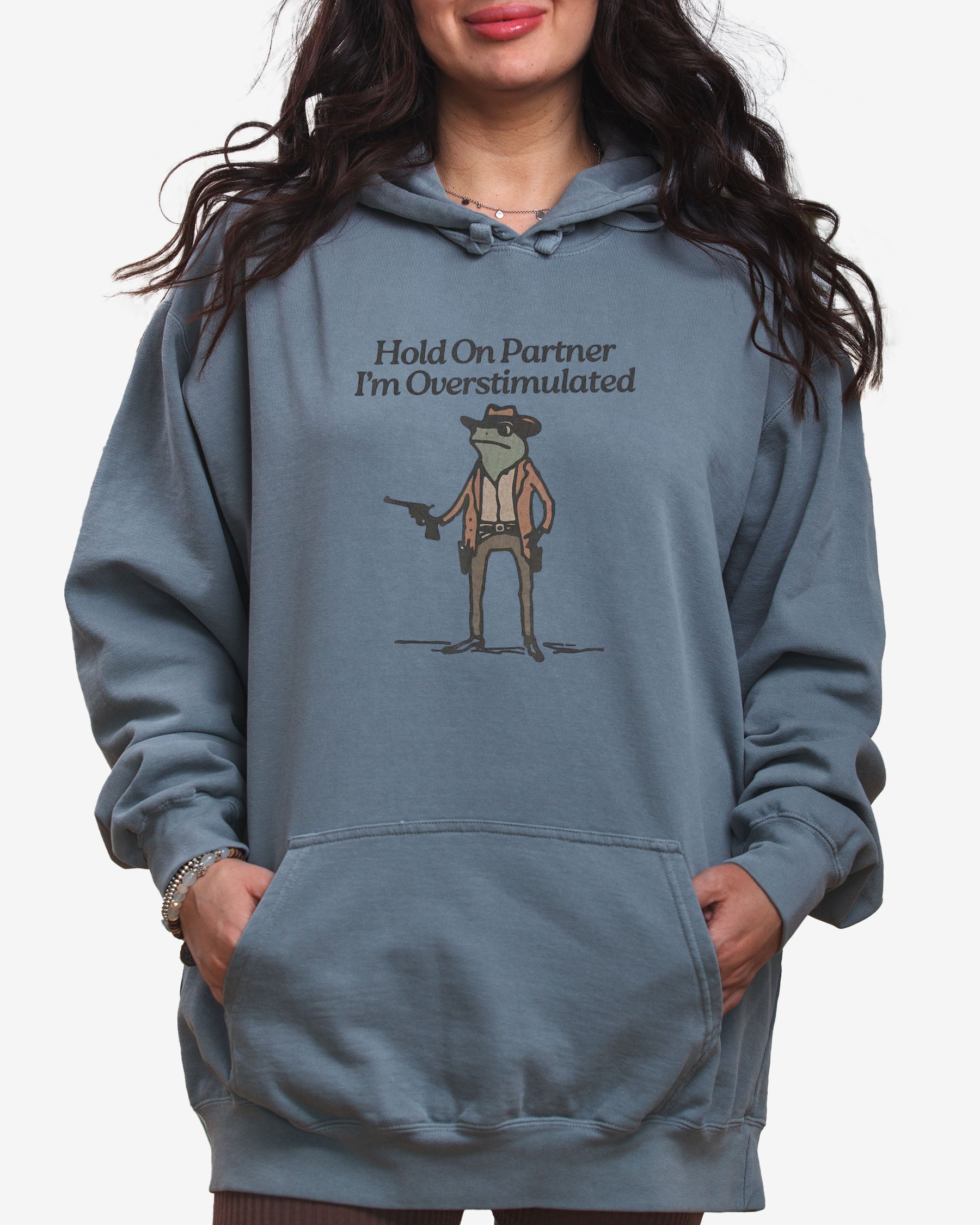 Woman wearing gray "Hold On Partner I'm Overstimulated" hoodie with a funny retro graphic of a cowboy frog.