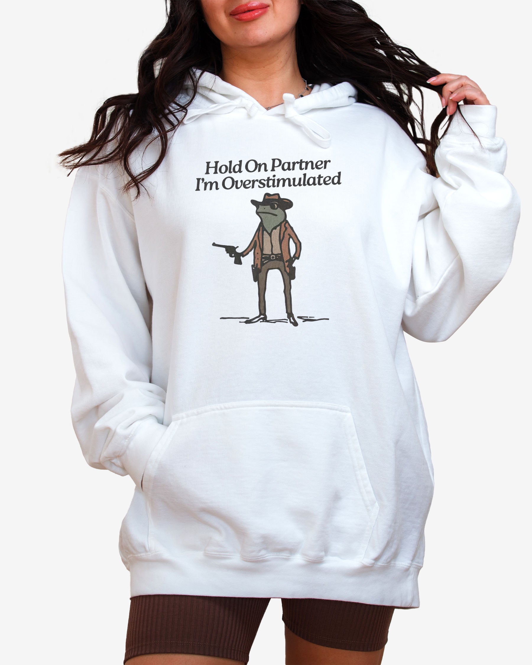 Woman wearing white "Hold On Partner I'm Overstimulated" hoodie with a funny retro graphic of a cowboy frog.