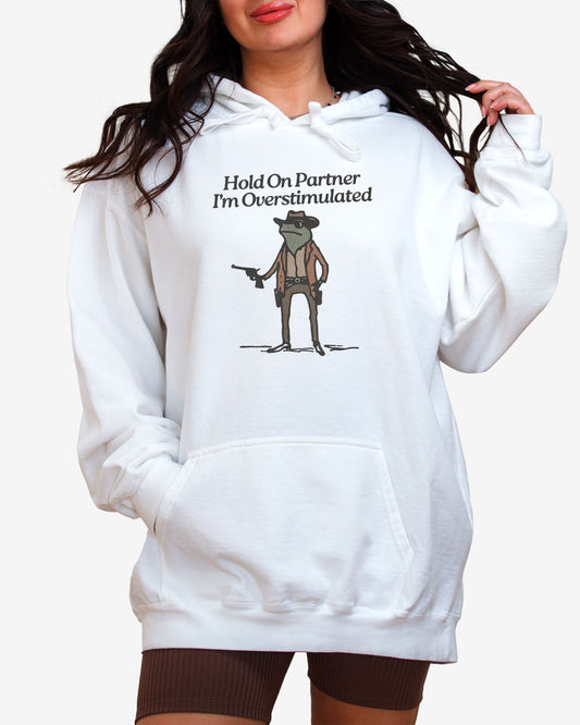 Woman wearing white "Hold On Partner I'm Overstimulated" hoodie with a funny retro graphic of a cowboy frog.