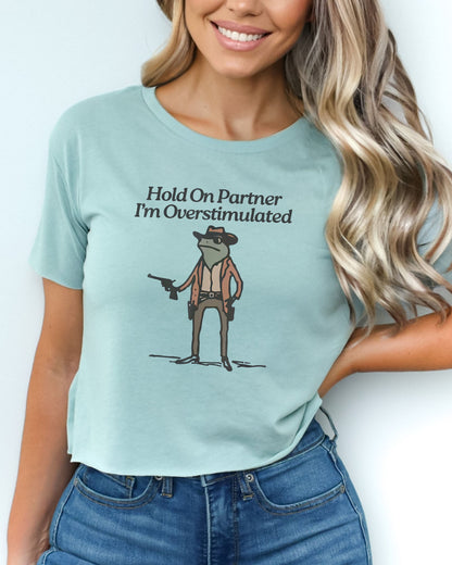 Woman wearing stonewash green 'Hold On Partner, I'm Overstimulated' crop top featuring a funny retro cowboy frog graphic.