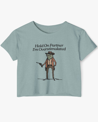 Flat lay stonewash green 'Hold On Partner, I'm Overstimulated' crop top featuring a funny retro cowboy frog graphic.