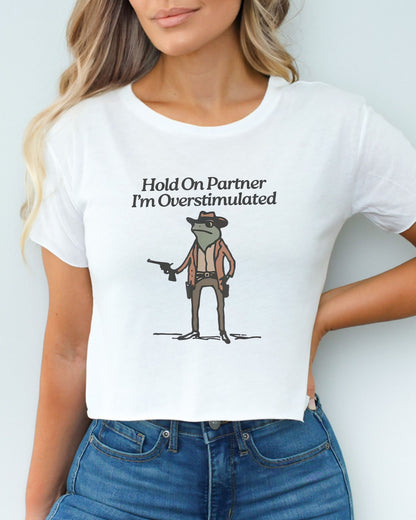 Woman wearing white 'Hold On Partner, I'm Overstimulated' crop top featuring a funny retro cowboy frog graphic.