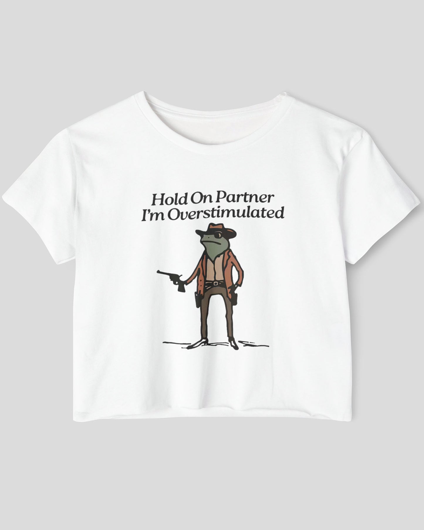 Flat lay white 'Hold On Partner, I'm Overstimulated' crop top featuring a funny retro cowboy frog graphic.