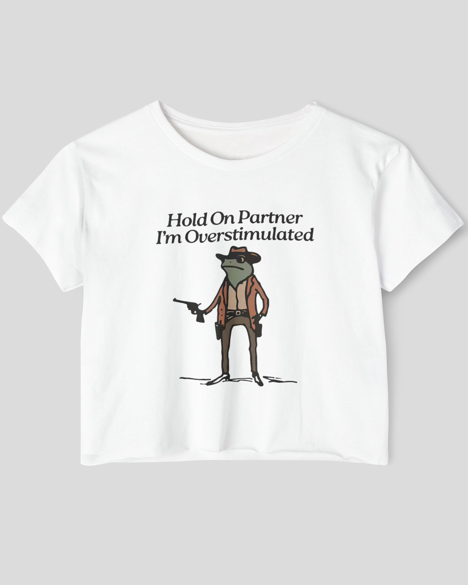Flat lay white 'Hold On Partner, I'm Overstimulated' crop top featuring a funny retro cowboy frog graphic.
