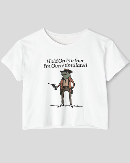 Flat lay white 'Hold On Partner, I'm Overstimulated' crop top featuring a funny retro cowboy frog graphic.