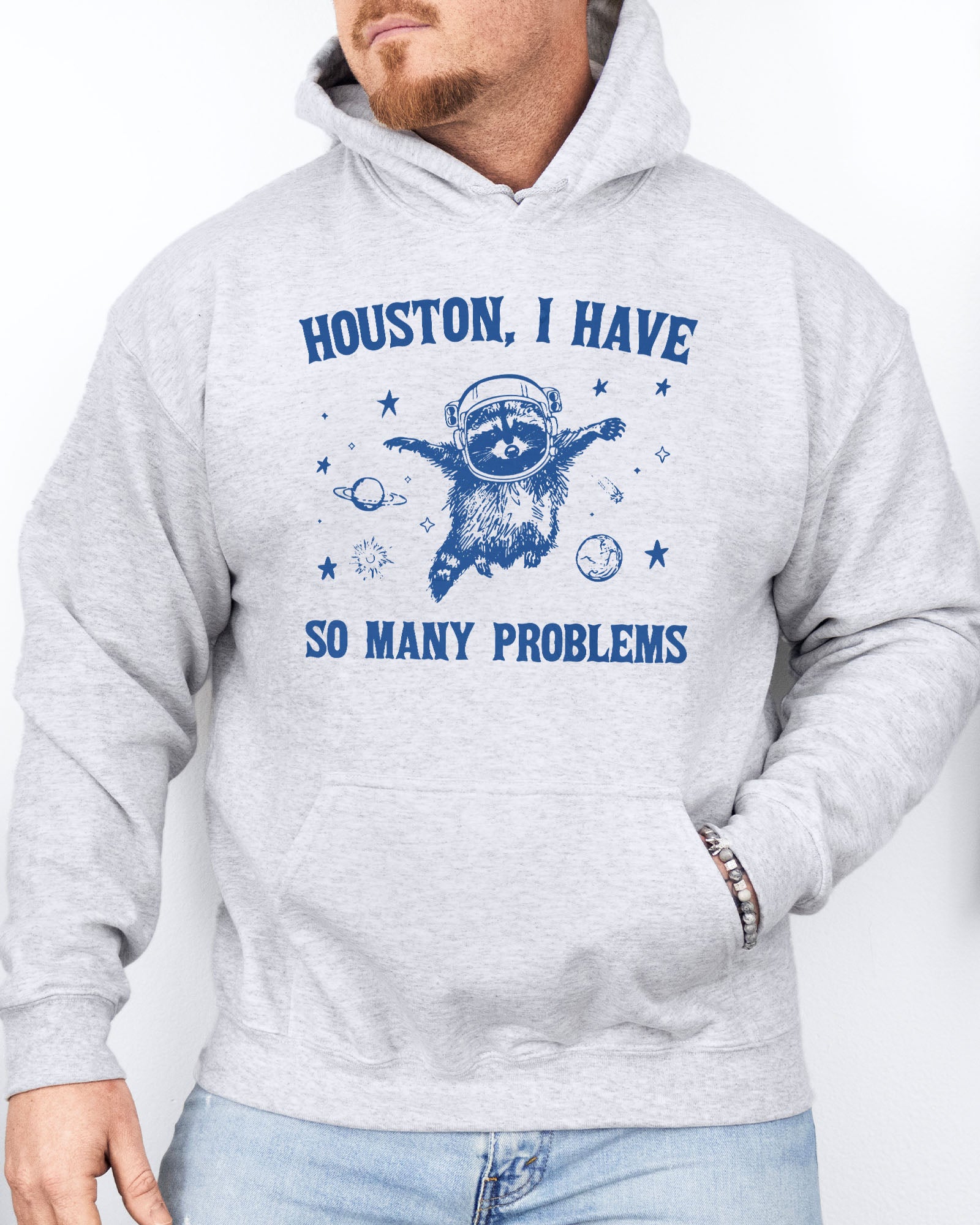 Man wearing an ash gray hoodie featuring a funny retro-style astronaut raccoon space graphic and the quote 'Houston I Have So Many Problems'.
