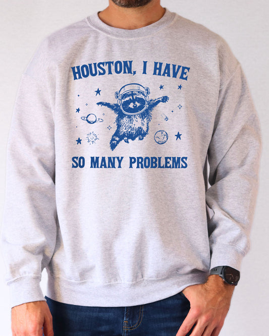Man wearing an ash gray sweatshirt featuring a funny retro astronaut raccoon graphic and the quote 'Houston I Have So Many Problems'.