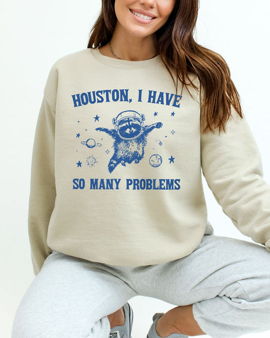 Woman wearing a sand beige sweatshirt featuring a funny retro astronaut raccoon graphic and the quote 'Houston I Have So Many Problems'.