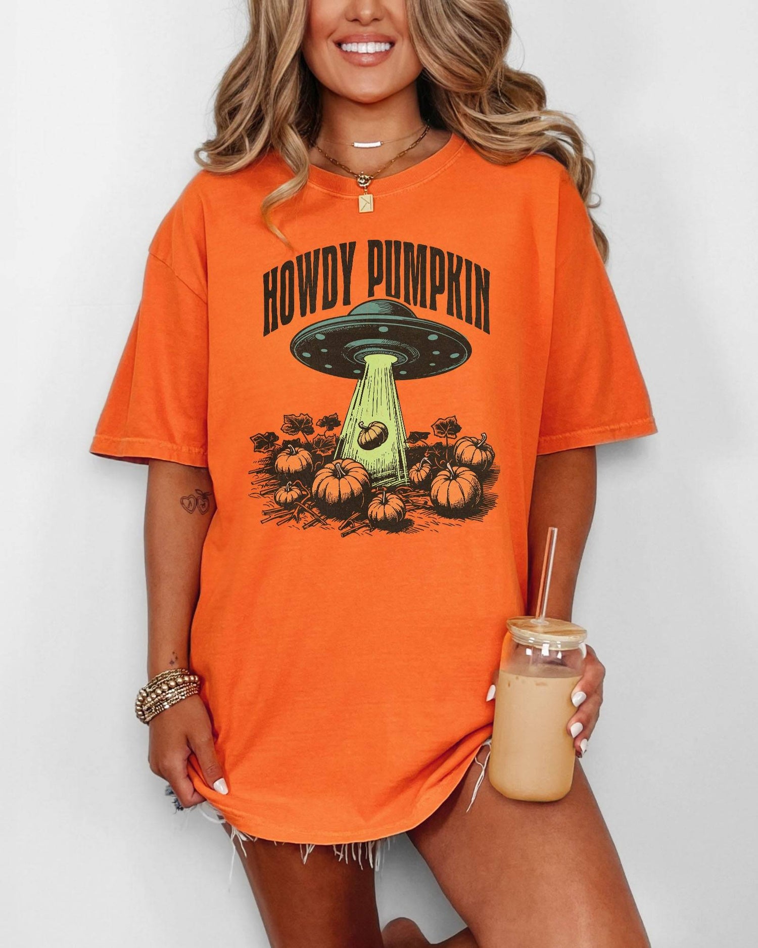 Woman wearing a burnt orange "Howdy Pumpkin" shirt with a retro graphic of a UFO abducting pumpkins from a field.