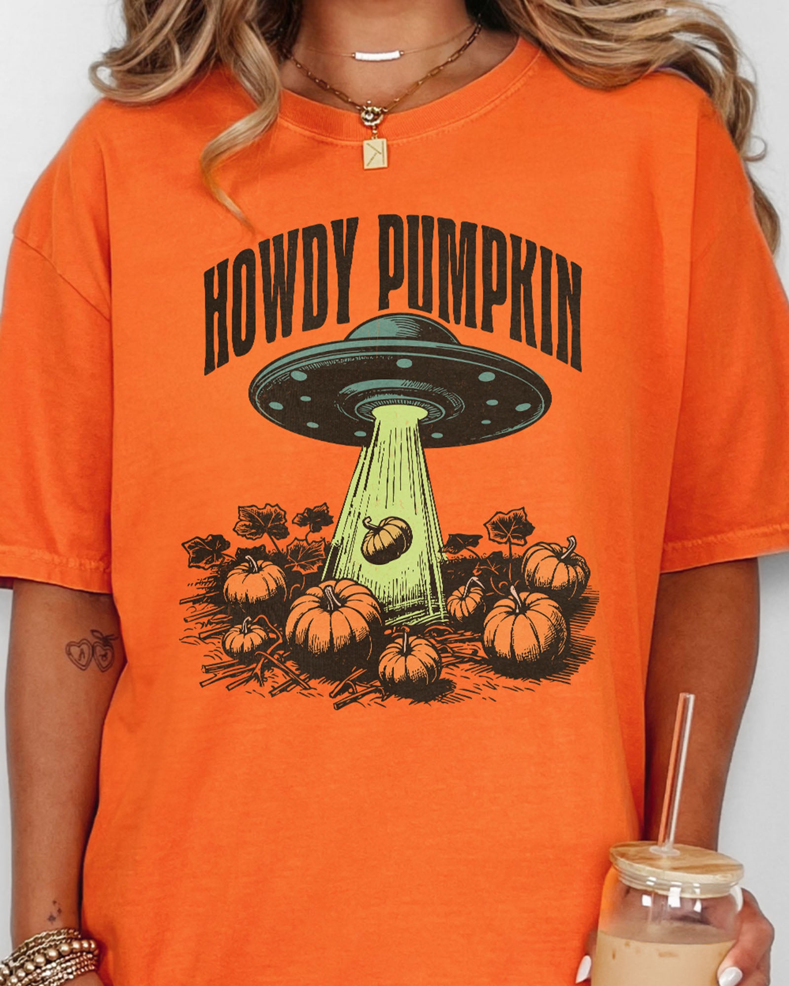 Close up of woman wearing a burnt orange "Howdy Pumpkin" shirt with a retro graphic of a UFO abducting pumpkins from a field.