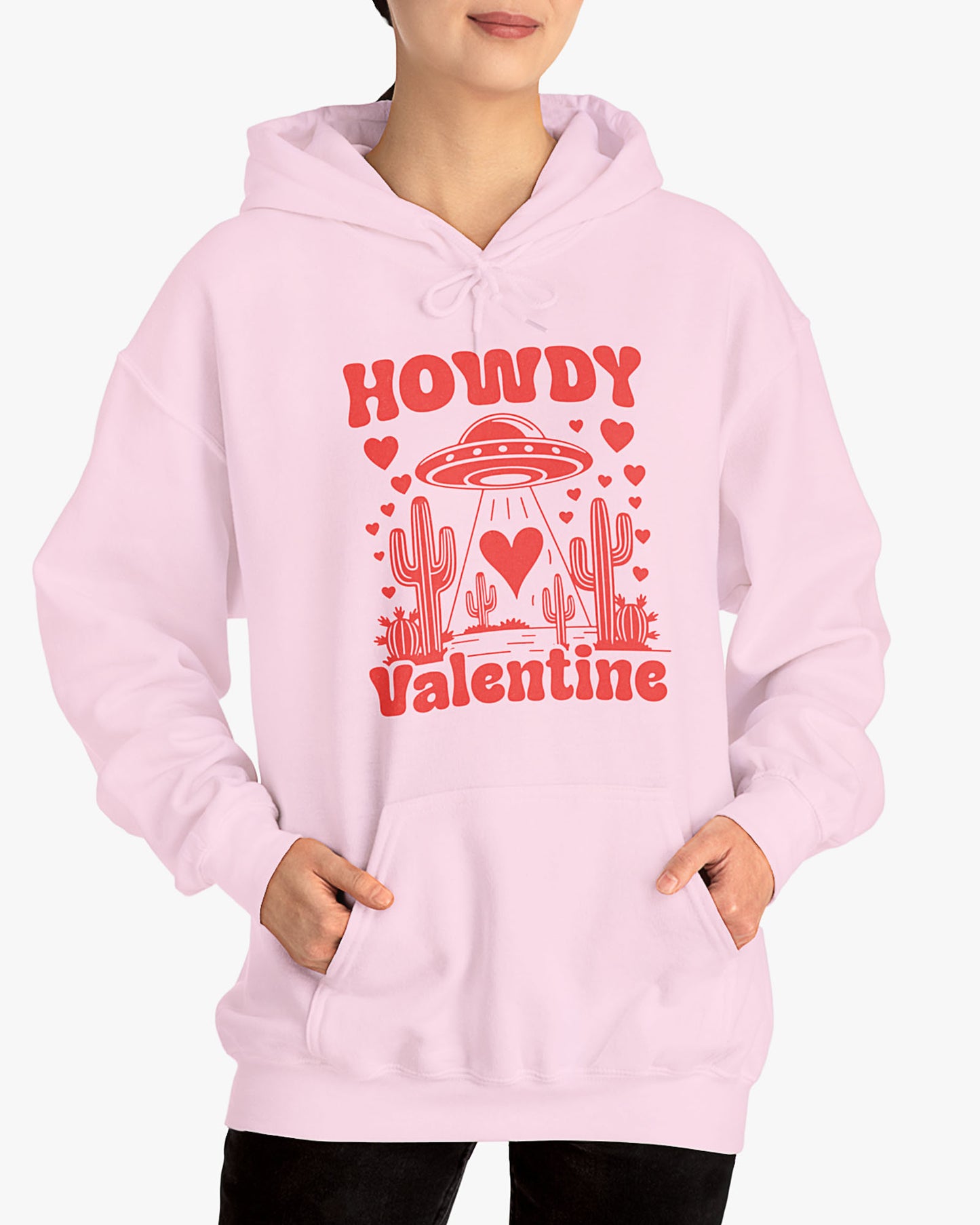 Woman wearing a light pink Howdy Valentine hoodie featuring a retro western desert scene with a UFO and floating hearts graphic.