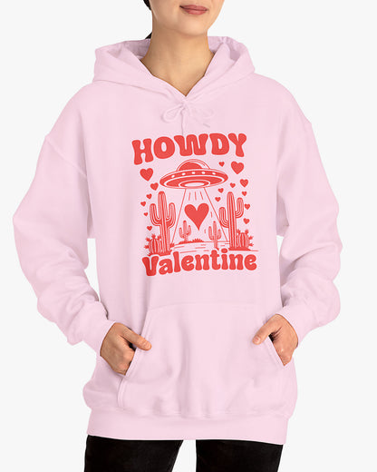 Woman wearing a light pink Howdy Valentine hoodie featuring a retro western desert scene with a UFO and floating hearts graphic.