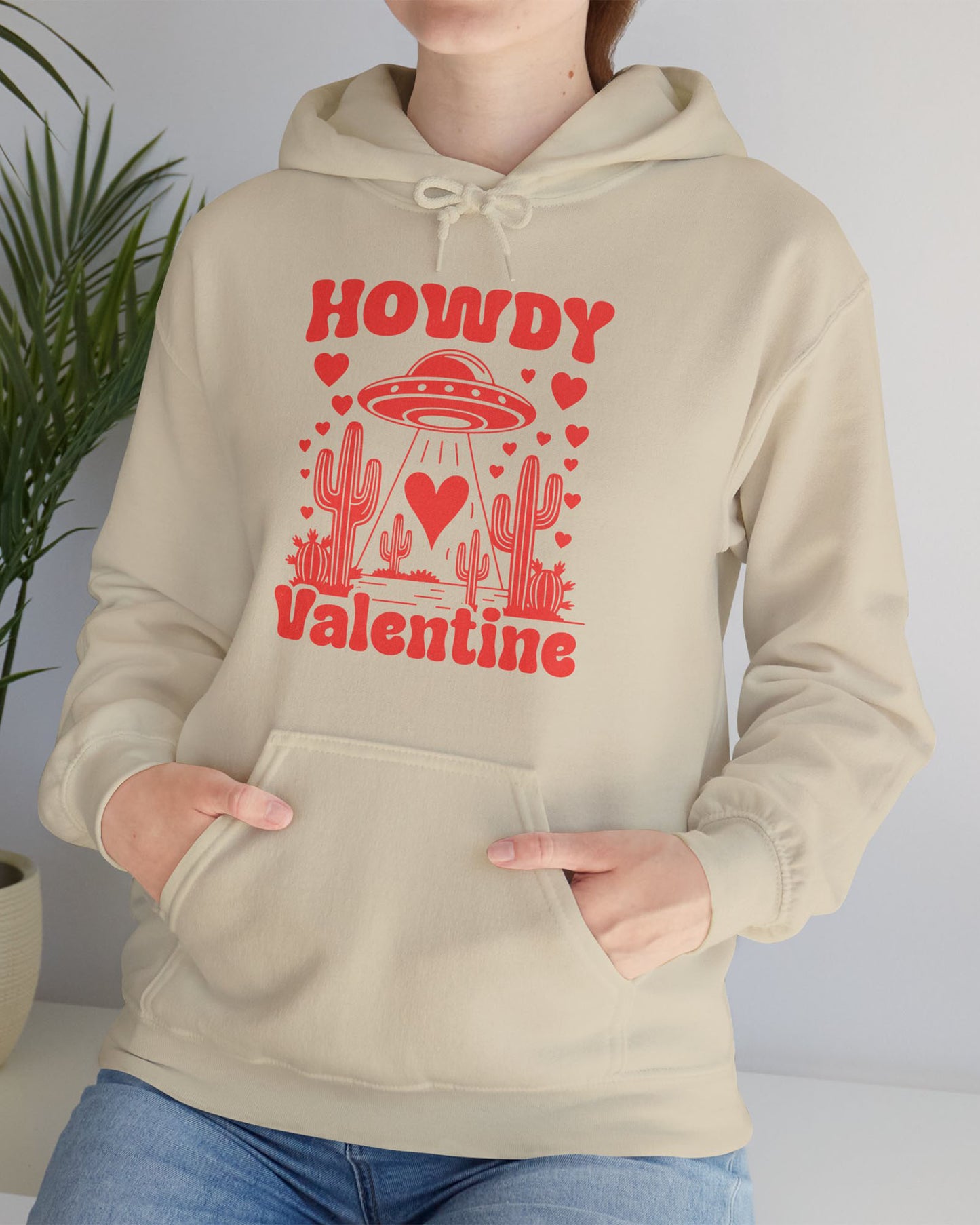 Woman wearing a sand beige Howdy Valentine hoodie featuring a retro western desert scene with a UFO and floating hearts graphic.