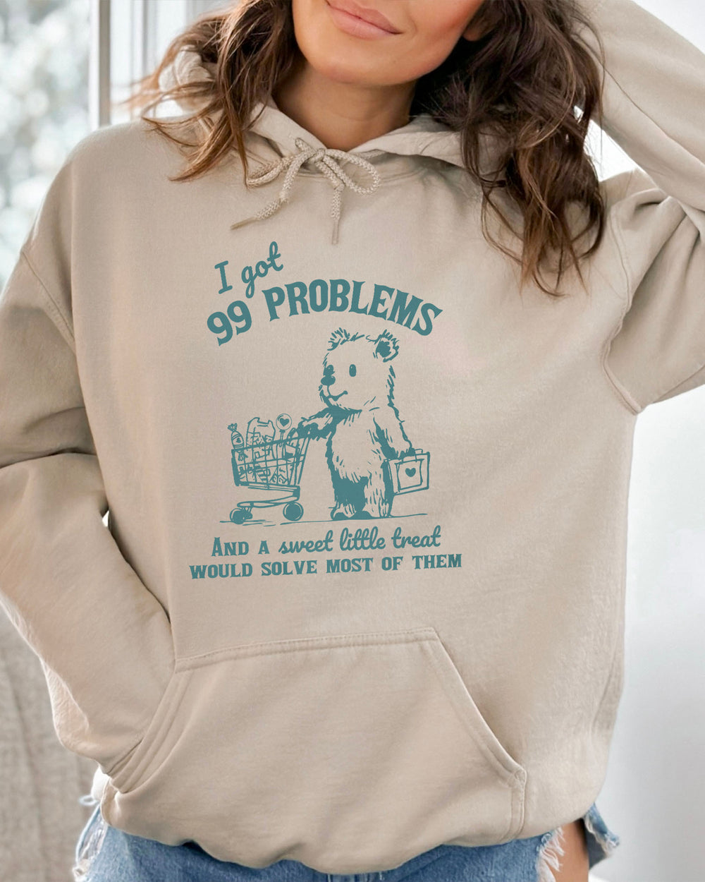 Woman wearing a sand beige hoodie with a cute, funny retro bear pushing a shopping cart and the quote 'I Got 99 Problems But A Sweet Little Treat Is Solving Most Of Them'.