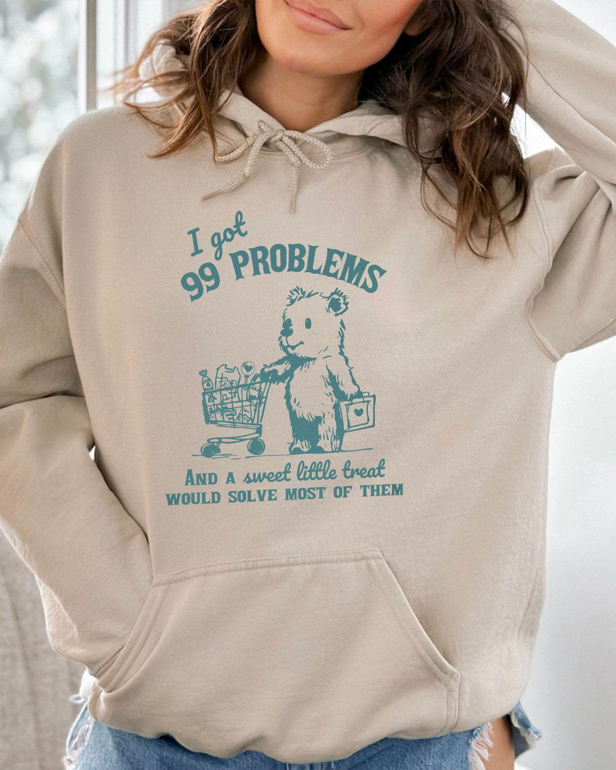 Woman wearing a sand beige hoodie with a cute, funny retro bear pushing a shopping cart and the quote 'I Got 99 Problems But A Sweet Little Treat Is Solving Most Of Them'.