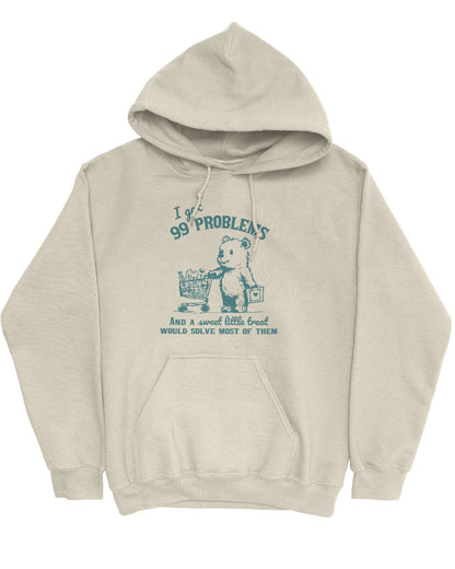 Flat lay of a sand beige hoodie with a cute, funny retro bear pushing a shopping cart and the quote 'I Got 99 Problems But A Sweet Little Treat Is Solving Most Of Them'.
