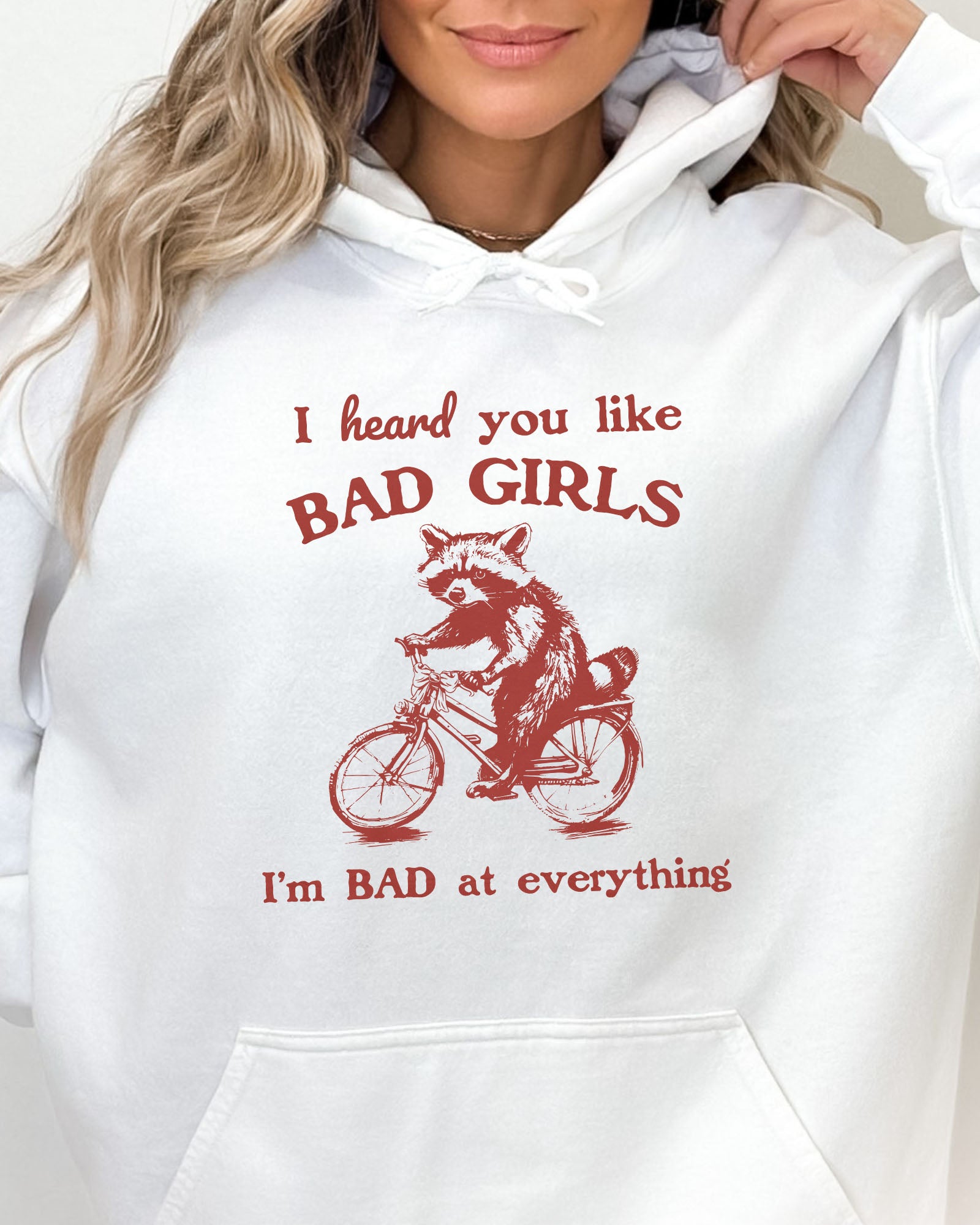 Close-up of a woman wearing a light pink hoodie featuring a funny retro-style raccoon riding a bycicle graphic and the quote 'I Heard You Like Bad Girls, I Am Bad At Everything'.