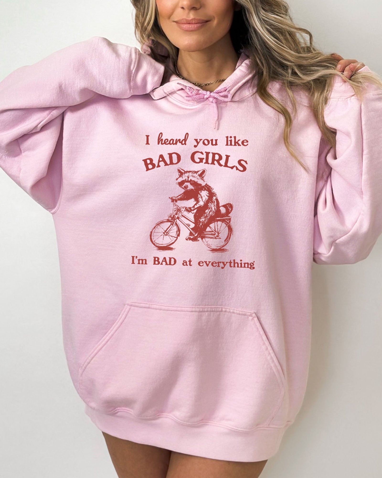 Woman wearing a light pink hoodie featuring a funny retro-style raccoon riding a bycicle graphic and the quote 'I Heard You Like Bad Girls, I Am Bad At Everything'.