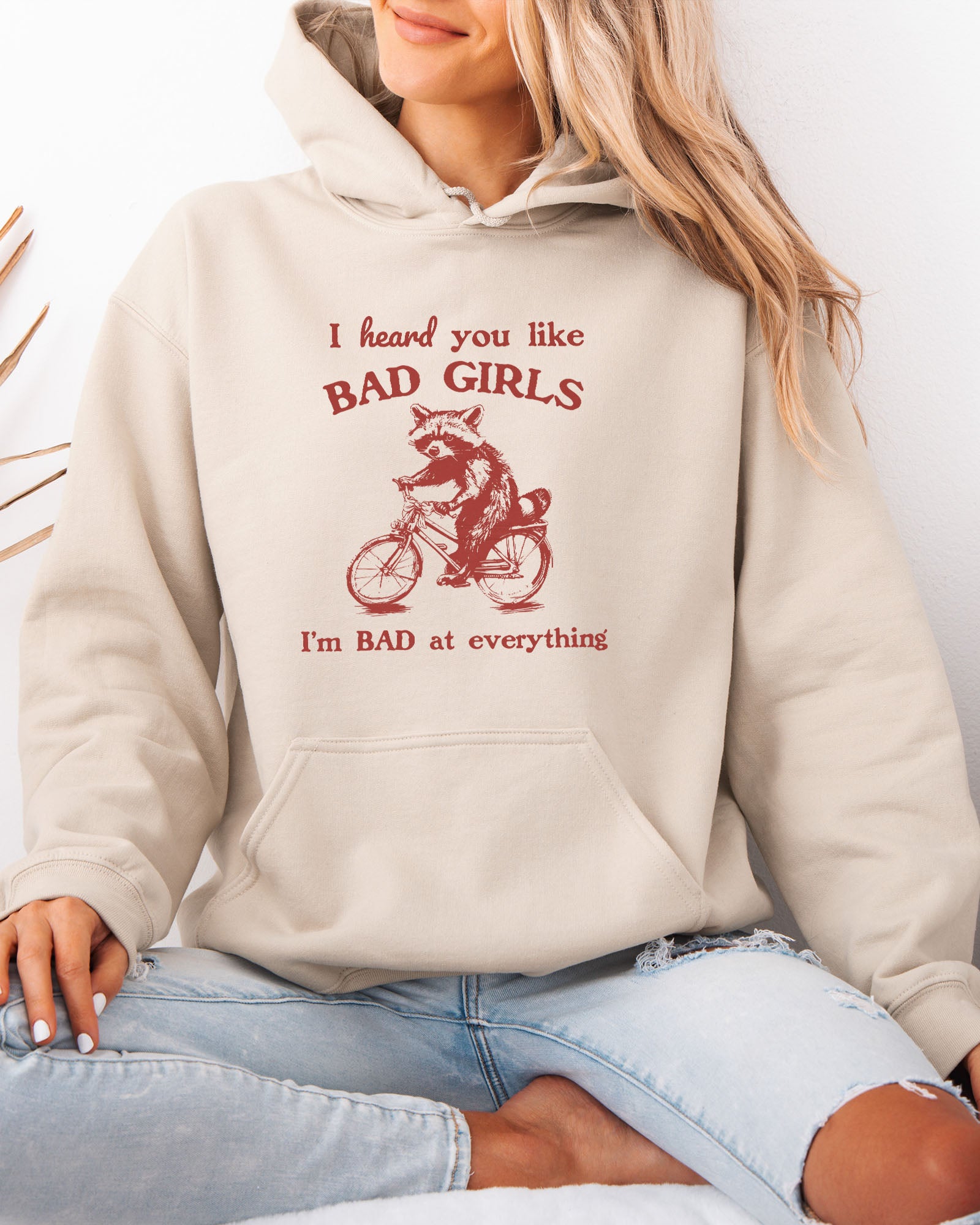 Woman wearing a sand beige hoodie featuring a funny retro-style raccoon riding a bycicle graphic and the quote 'I Heard You Like Bad Girls, I Am Bad At Everything'.